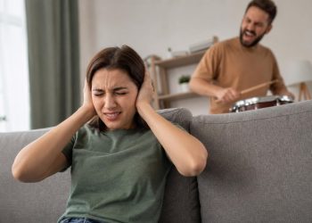 Distressed woman covering her ears on a couch while a man plays loud drums and shouts in the background.