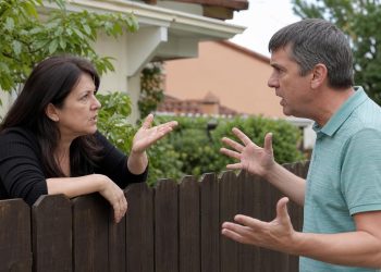 A man and woman having a heated argument with expressive hand gestures over a dark wooden garden fence between their houses.