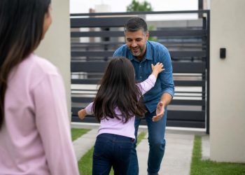 Smiling father in a denim shirt opens his arms to greet his young daughter as she runs toward him, while the mother watches from the foreground.