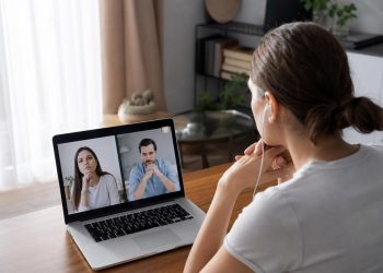 Woman with earphones at a desk hosting a video call on her laptop with a man and woman who appear to be in a serious discussion.